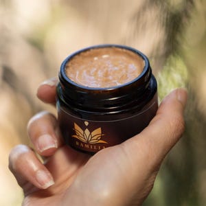 May include: A small, dark brown jar of skincare product held in a hand. The jar has a gold label with a lotus flower design and the word "RAMILIA". The product inside is a light brown color.