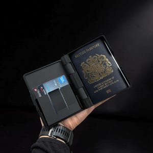 May include: Black passport holder with a British passport and a credit card. The passport is dark blue with gold lettering and a crest. The holder has a slot for the passport and a slot for a credit card.