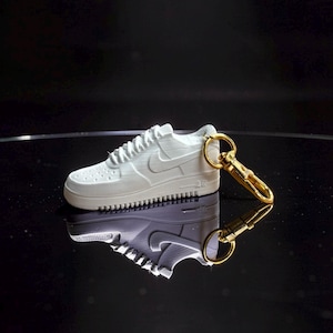 Può includere: Un portachiavi a forma di sneaker bianca in miniatura con chiusura color oro. La sneaker ha una superficie strutturata, un logo Nike e una suola dettagliata. Il portachiavi si riflette su una superficie nera lucida.