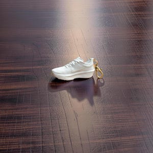 Può includere: Un portachiavi a forma di sneaker bianca con chiusura color oro. La scarpa in miniatura presenta una suola strutturata e un design a virgola sul lato. Il portachiavi è una replica in miniatura di una scarpa da corsa.