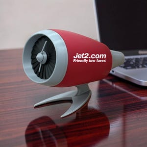 Puede incluir: Altavoz de escritorio rojo y gris con forma de motor a reacción. El altavoz tiene un diseño similar a un ventilador en la parte delantera y el texto "Jet2.com Friendly low fares" en el lateral. Se encuentra sobre una superficie de madera.