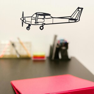 May include: Black metal airplane wall art. The airplane features a propeller, windows, and tail. The art is mounted on a white wall above a desk with a red binder and office supplies.