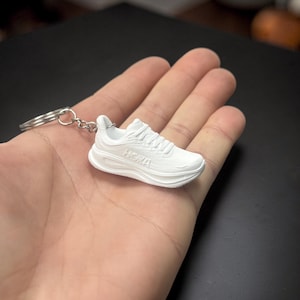 May include: A white miniature sneaker keychain with the brand name "HOKA" visible on the side. The keychain is made of a solid white material and includes a silver-colored keyring.
