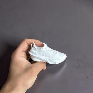 May include: A small, white sneaker with intricate detailing, held in a hand. The miniature shoe features a textured sole, laces, and a curved design, showcasing a modern athletic style. The shoe is a solid white color.