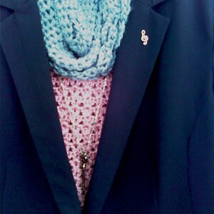 May include: A navy blue blazer with a light blue crocheted infinity scarf and a pink crocheted top. A silver-coloured treble clef pin is on the blazer. A silver necklace with a key pendant is visible.