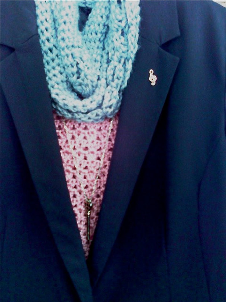 May include: A navy blue blazer with a light blue crocheted infinity scarf and a pink crocheted top. A silver-coloured treble clef pin is on the blazer. A silver necklace with a key pendant is visible.