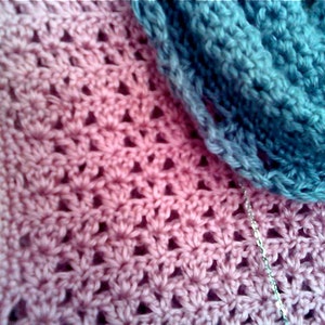 May include: Close-up of a handmade crochet item in shades of pink and blue. The pink section features a floral pattern, while the blue section appears to be a scarf or cowl. A silver chain is visible, adding a touch of detail to the textile.