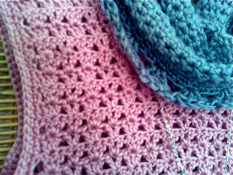 May include: Close-up of a handmade crochet item in shades of pink and blue. The pink section features a floral pattern, while the blue section appears to be a scarf or cowl. A silver chain is visible, adding a touch of detail to the textile.