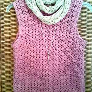 May include: A sleeveless, dusky pink crochet top with a matching crochet scarf and a silver pendant necklace. The top is displayed on a clear hanger. The scarf is a light beige colour. The crochet pattern is visible.