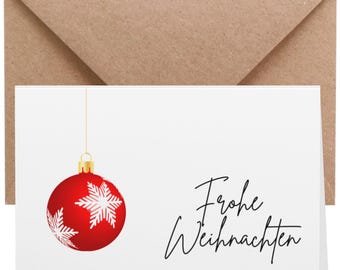 Könnte beinhalten: Weihnachtskarte mit roter Christbaumkugel, verziert mit weißen Schneeflocken und dem Schriftzug "Frohe Weihnachten" in schwarzer Schrift. Die Karte liegt vor einem weißen Hintergrund, mit einem braunen Umschlag.