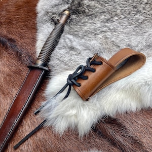 May include: A brown leather knife sheath with decorative lacing detail. The sheath is lying on a brown and white fur surface. A brown leather knife with a brass handle is also visible in the image.