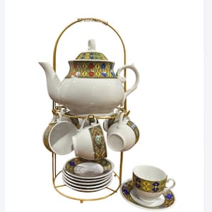 May include: A white ceramic tea set on a gold-coloured metal stand. The set includes a teapot, six cups, and saucers. The teapot and cups feature a colourful geometric pattern in yellow, blue, and red. The stand has a handle for easy carrying.