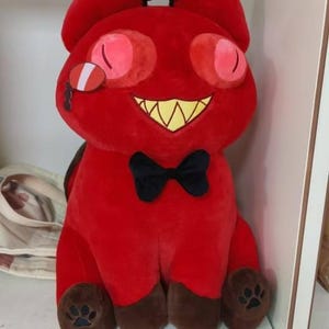 May include: A plush red cat-like character with brown ears and paws, a black bow tie, and a wide, toothy grin. The character has large pink eyes and a small, white and red detail on its cheek. The plush is sitting on a shelf.