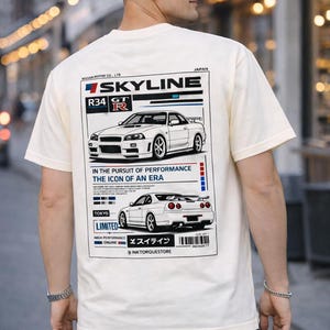 Può includere: T-shirt color crema con grafica di un'auto Nissan Skyline R34 GT-R bianca. Il design include il testo "SKYLINE", "R34 GT-R" e "IN THE PURSUIT OF PERFORMANCE THE ICON OF AN ERA". La maglietta ha girocollo e maniche corte.