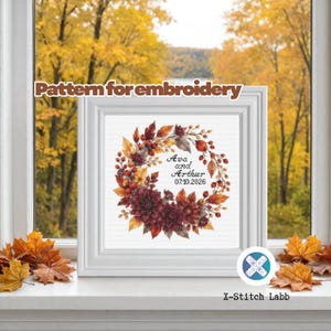 May include: A white square frame containing an embroidery pattern of an autumn wreath with red and orange leaves, berries, and dark flowers. The text "Ava and Arthur 07.10.2026" is centred within the wreath. The text "Pattern for embroidery" appears above.