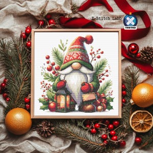 Könnte beinhalten: Ein gerahmtes Kreuzstichbild mit einem Weihnachtszwerg mit langem weißem Bart, roter Mütze und grünem Mantel, der eine Laterne hält. Das Kunstwerk ist von festlichem Grün, roten Beeren und Orangen umgeben.