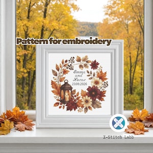 May include: A square white frame containing an embroidery pattern of an autumn wreath with red, orange, and brown leaves, berries, and a lantern. The centre features the text Emma and Lucas 23.09.2026. The text Pattern for embroidery is visible.