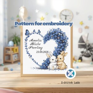 Peut inclure: Un modèle de broderie représentant une couronne en forme de cœur faite de fleurs bleues et de papillons, avec le texte 'Amelia Blake Sterling' et '23.09.2026'. Deux petits lapins, un blanc et un beige, sont assis au pied du motif floral.