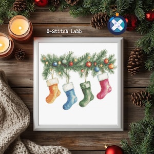 Op de afbeelding: Een ingelijste kruissteek met vier kleurrijke kerstsokken die aan een slinger met ornamenten hangen. De sokken zijn oranje, blauw, groen en roze, elk met een witte manchet. De tekst "X-Stitch Labb" is zichtbaar.