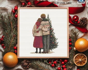 May include: Cross-stitch art of a couple embracing near a Christmas tree. The artwork is in a wooden frame, surrounded by festive decorations like pine branches, red berries, and an orange. The text "X-Stitch Labb" is visible.