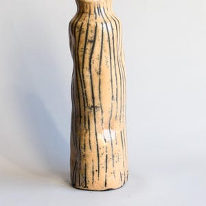 May include: A tall, handmade ceramic vase with a textured surface. The vase is a light tan color with vertical black lines and a slightly irregular shape. The vase has a narrow neck and is suitable for home decor.