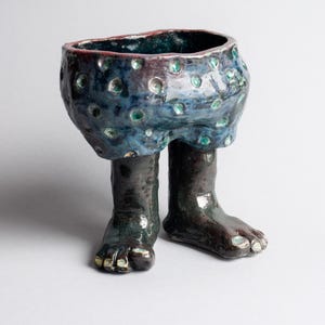 May include: A whimsical ceramic bowl with a blue, textured exterior adorned with green and maroon accents. The bowl is supported by two dark brown legs and feet, creating a unique, artistic design.