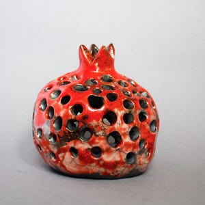 May include: A red ceramic pomegranate with a textured surface and a hollow interior. The pomegranate has a small crown on top and is decorated with black and white accents.