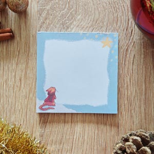 May include: A square, light blue sticky note with a white center and a light blue border. A small illustration of a character in a red hat is in the corner. A yellow star is in the corner. The note is on a wooden surface with Christmas decorations.