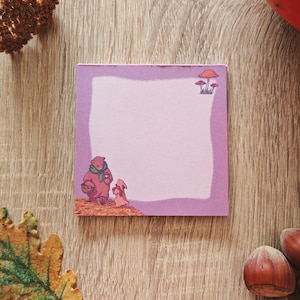 Peut inclure: Un bloc-notes carré de couleur lavande avec une illustration fantaisiste d'un ours et d'un lapin, ainsi que des champignons. Le bloc-notes a un intérieur rose clair et une bordure décorative. Le fond est une surface en bois avec des éléments d'automne.