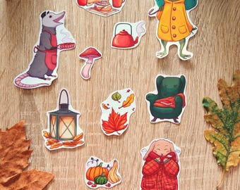 Op de afbeelding: Een verzameling herfststickers met illustraties van een opossum met taart, een kikker met paraplu, een konijn in een geruite jas, pompoenen, een paddenstoel, een theepot en een lantaarn. De stickers liggen op een houten oppervlak.