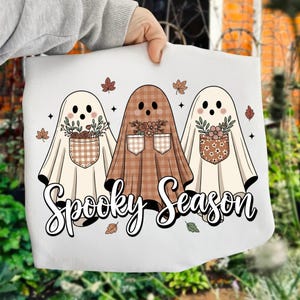 May include: A white tote bag featuring three cartoon ghosts with floral pocket details and the text "Spooky Season". The ghosts are in cream and brown tones, with autumn leaves scattered around.
