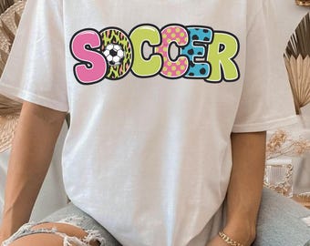 May include: White t-shirt with the word "SOCCER" in colorful, bold letters. The letter "S" is pink, the "O" has a soccer ball design, the "C" is lime green, the second "C" has pink polka dots, the "E" is blue, and the "R" is green.