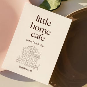Puede incluir: Una tarjeta color crema con las palabras "little home café" en letras marrones. Debajo, se lee "coffee, bites & vibes". Un dibujo lineal de un café con el texto "Sophie's Café" está en la parte inferior.