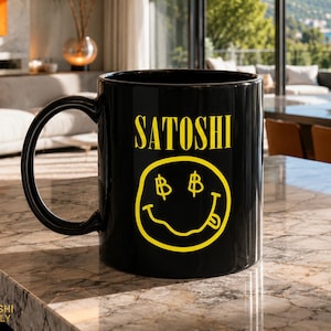 Op de afbeelding: Zwarte keramische mok met het woord "SATOSHI" in geel, boven een gele smiley met dollartekenogen en een uitgestoken tong. De mok heeft een grote zwarte handgreep en staat op een marmeren aanrecht.