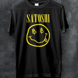 Peut inclure: T-shirt noir avec le mot "Satoshi" en jaune au-dessus d'un smiley jaune avec des symboles dollar pour les yeux et une langue tir&eacute;e. Le t-shirt est suspendu &agrave; un cintre blanc.