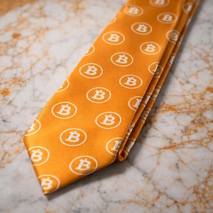 Könnte beinhalten: Eine orangefarbene Krawatte mit einem sich wiederholenden Muster aus weißen Bitcoin-Symbolen. Die Krawatte ist gefaltet und liegt auf einer Marmoroberfläche. Die Bitcoin-Symbole befinden sich in weißen Kreisen.