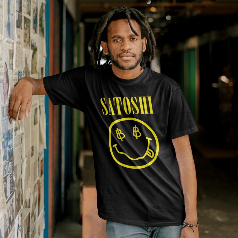 Peut inclure: T-shirt noir avec le mot "SATOSHI" en jaune au-dessus d'un smiley jaune avec des yeux en forme de symbole dollar et une langue tir&eacute;e. Le t-shirt est &agrave; manches courtes.