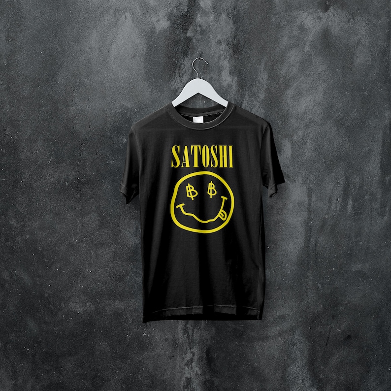 Peut inclure: T-shirt noir avec le mot "Satoshi" en jaune au-dessus d'un smiley jaune avec des symboles dollar pour les yeux et une langue tir&eacute;e. Le t-shirt est suspendu &agrave; un cintre blanc.