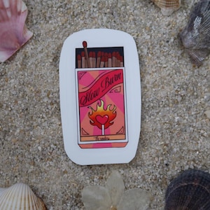 Peut inclure: Autocollant blanc représentant une boîte d'allumettes avec l'inscription "Slow Burn" en cursive. La boîte est rose et orange avec un motif de flamme en forme de cœur. L'autocollant est entouré de coquillages et de sable.