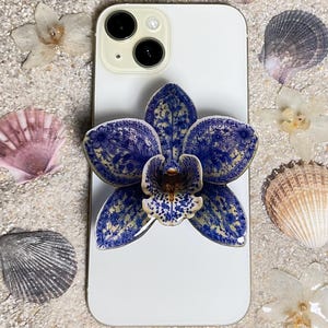 Peut inclure: Un smartphone blanc avec un support de téléphone à motif floral bleu et blanc. Le téléphone est entouré de coquillages et de fleurs séchées sur une surface sablonneuse. Le support de téléphone a la forme d'une orchidée.