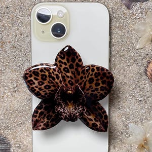 May include: A white smartphone with a leopard print flower phone grip. The flower grip has a brown and black spotted pattern. The phone is on a sandy surface with seashells and dried flowers.