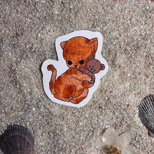 May include: A sticker featuring an orange cat holding a brown teddy bear. The cat has large black eyes and a white outline. The sticker is on a sandy surface with seashells and a flower.