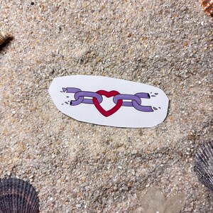 May include: A sticker featuring a broken chain design with a red heart in the center. The chain links are purple, and the heart is red. The sticker is on a white background, surrounded by sand and seashells.