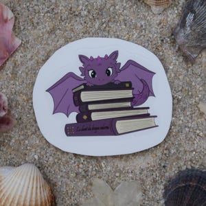 Peut inclure: Autocollant de dragon violet avec une pile de livres, entouré de coquillages et de fleurs sur une surface sablonneuse. Le dragon a de grandes ailes et est perché sur les livres. Les livres ont le texte "La vie est un livre ouvert".
