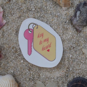 Peut inclure: Un autocollant avec une clé rose et une étiquette avec l'inscription "key to my heart" et un petit cœur rouge. L'autocollant est entouré de coquillages et d'une fleur sur une surface sablonneuse.