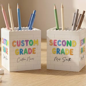May include: Two white square pencil holders constructed from interlocking building blocks. The left holder features the text "CUSTOM GRADE" and "Custom Name" in colorful letters. The right holder displays "SECOND GRADE" and "Mrs. Smith".