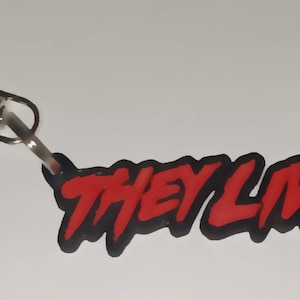 Könnte beinhalten: Ein roter und schwarzer Schlüsselanhänger mit den Worten "They Live" in einer stilisierten Schriftart. Der Schlüsselanhänger hat einen silbernen Metallclip.