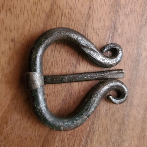 May include: Hand-forged iron buckle with a distinctive scroll design. The buckle features a curved body with a pin closure. The metal has a dark, textured finish, suggesting a rustic or antique aesthetic. The buckle is set against a wooden background.