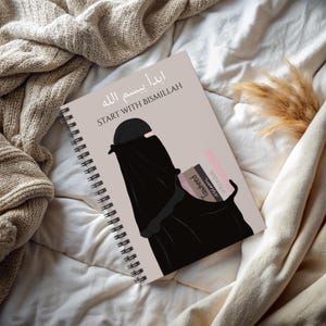 May include: Spiral-bound notebook with a beige cover featuring an illustration of a woman in a black niqab. The notebook has the text "START WITH BISMILLAH" and Arabic script. The background is a neutral beige.