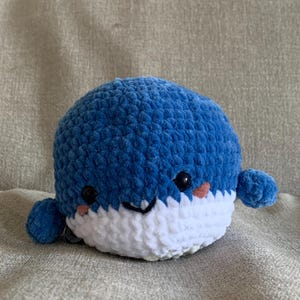 May include: A blue and white crocheted whale plush toy with a smiling face. The whale has small black eyes and pink cheeks.